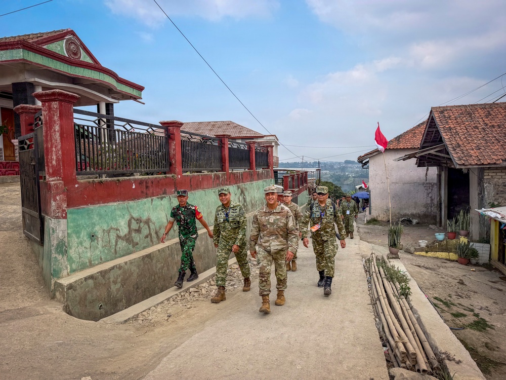 A group of military personnel in camouflage uniforms are walking together along a concrete path through a rural village. The group appears to include soldiers from at least two different military forces, identifiable by slight uniform variations. A red and white flag, consistent with the Indonesian national flag, is visible in the background, suggesting this is located in Indonesia.