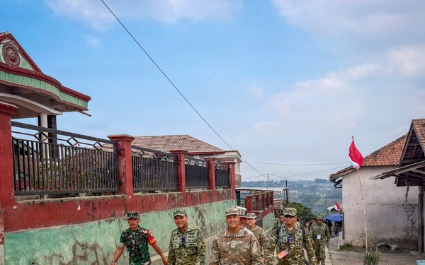 A group of military personnel in camouflage uniforms are walking together along a concrete path through a rural village. The group appears to include soldiers from at least two different military forces, identifiable by slight uniform variations. A red and white flag, consistent with the Indonesian national flag, is visible in the background, suggesting this is located in Indonesia.