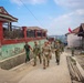 A group of military personnel in camouflage uniforms are walking together along a concrete path through a rural village. The group appears to include soldiers from at least two different military forces, identifiable by slight uniform variations. A red and white flag, consistent with the Indonesian national flag, is visible in the background, suggesting this is located in Indonesia.