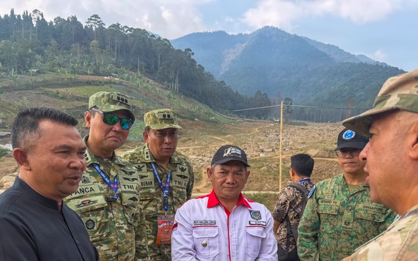 A group of military and civilian personnel are gathered outdoors in a mountainous, partially deforested area, engaged in what appears to be a briefing or discussion. The group includes soldiers wearing TNI (Indonesian Army) uniforms, a Singapore Armed Forces member, a US Army soldier in OCP camouflage, and civilian officials. The setting suggests a disaster response assessment or joint military exercise site visit.