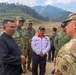 A group of military and civilian personnel are gathered outdoors in a mountainous, partially deforested area, engaged in what appears to be a briefing or discussion. The group includes soldiers wearing TNI (Indonesian Army) uniforms, a Singapore Armed Forces member, a US Army soldier in OCP camouflage, and civilian officials. The setting suggests a disaster response assessment or joint military exercise site visit.