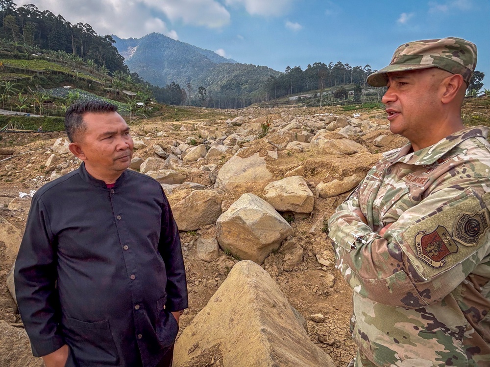 Two men are standing and conversing at what appears to be a landslide or debris field site, with large boulders and rocky debris spread across a wide area. One man is wearing civilian clothing (black jacket) while the other is in military camouflage uniform with patches and a cap. The background shows forested mountains and a valley filled with displaced rocks and sediment, consistent with a natural disaster assessment or relief operation.