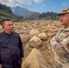 Two men are standing and conversing at what appears to be a landslide or debris field site, with large boulders and rocky debris spread across a wide area. One man is wearing civilian clothing (black jacket) while the other is in military camouflage uniform with patches and a cap. The background shows forested mountains and a valley filled with displaced rocks and sediment, consistent with a natural disaster assessment or relief operation.