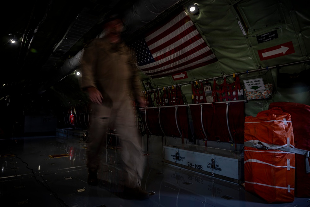 The interior of a military transport aircraft, likely a C-130 or similar cargo plane, is shown in low-light conditions. A motion-blurred figure in a tan/khaki flight suit is moving through the cargo bay. The aircraft interior features an American flag, safety signage, red cargo bags, and various military equipment mounted along the fuselage walls.