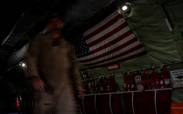 The interior of a military transport aircraft, likely a C-130 or similar cargo plane, is shown in low-light conditions. A motion-blurred figure in a tan/khaki flight suit is moving through the cargo bay. The aircraft interior features an American flag, safety signage, red cargo bags, and various military equipment mounted along the fuselage walls.