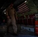 The interior of a military transport aircraft, likely a C-130 or similar cargo plane, is shown in low-light conditions. A motion-blurred figure in a tan/khaki flight suit is moving through the cargo bay. The aircraft interior features an American flag, safety signage, red cargo bags, and various military equipment mounted along the fuselage walls.