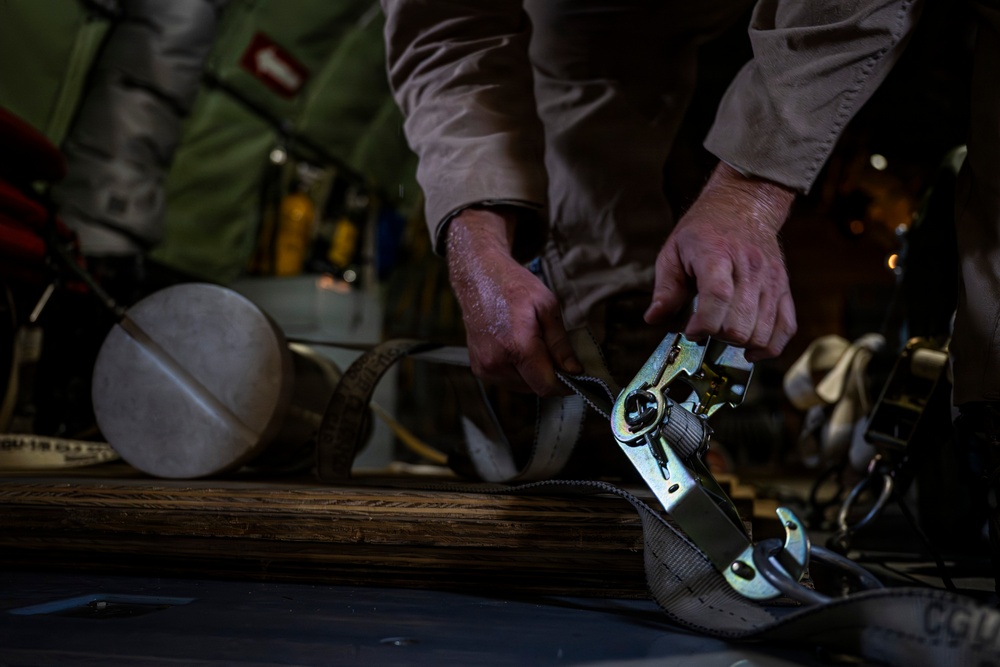 A person in tan/khaki clothing is working with ratchet straps to secure cargo, likely inside a military transport aircraft. The image shows hands manipulating a metal ratchet buckle attached to webbing straps over what appears to be wooden rollers or logs. The low-light, close-up composition emphasizes the hands and hardware in detail.