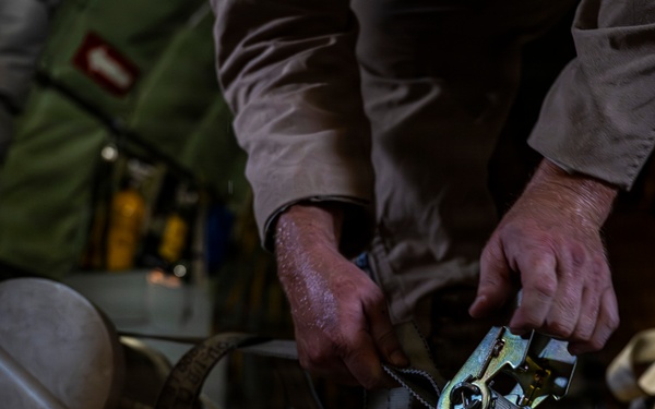 A person in tan/khaki clothing is working with ratchet straps to secure cargo, likely inside a military transport aircraft. The image shows hands manipulating a metal ratchet buckle attached to webbing straps over what appears to be wooden rollers or logs. The low-light, close-up composition emphasizes the hands and hardware in detail.