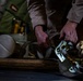 A person in tan/khaki clothing is working with ratchet straps to secure cargo, likely inside a military transport aircraft. The image shows hands manipulating a metal ratchet buckle attached to webbing straps over what appears to be wooden rollers or logs. The low-light, close-up composition emphasizes the hands and hardware in detail.
