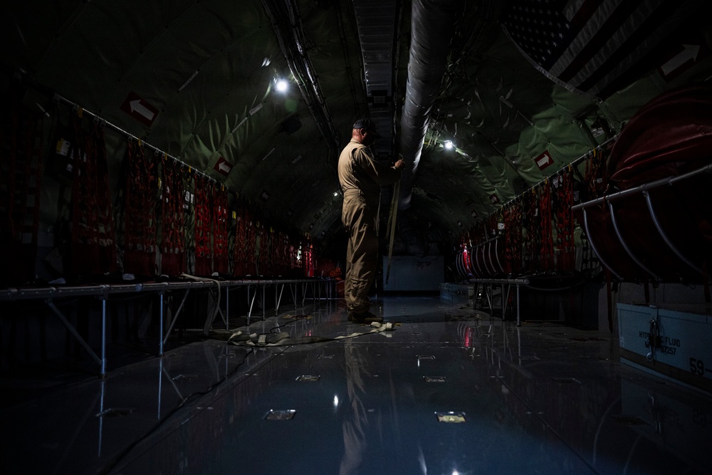 A person in military-style olive/tan clothing stands inside the cargo hold of a large military transport aircraft, appearing to inspect or handle overhead ducting or equipment. The interior is dimly lit with overhead lighting illuminating the long, tunnel-like fuselage. Red cargo tie-down straps and safety markings line both walls, and the reflective metal floor shows scattered straps and hardware.