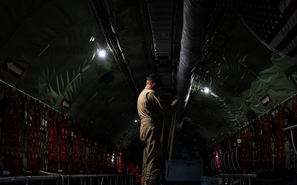 A person in military-style olive/tan clothing stands inside the cargo hold of a large military transport aircraft, appearing to inspect or handle overhead ducting or equipment. The interior is dimly lit with overhead lighting illuminating the long, tunnel-like fuselage. Red cargo tie-down straps and safety markings line both walls, and the reflective metal floor shows scattered straps and hardware.