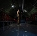A person in military-style olive/tan clothing stands inside the cargo hold of a large military transport aircraft, appearing to inspect or handle overhead ducting or equipment. The interior is dimly lit with overhead lighting illuminating the long, tunnel-like fuselage. Red cargo tie-down straps and safety markings line both walls, and the reflective metal floor shows scattered straps and hardware.