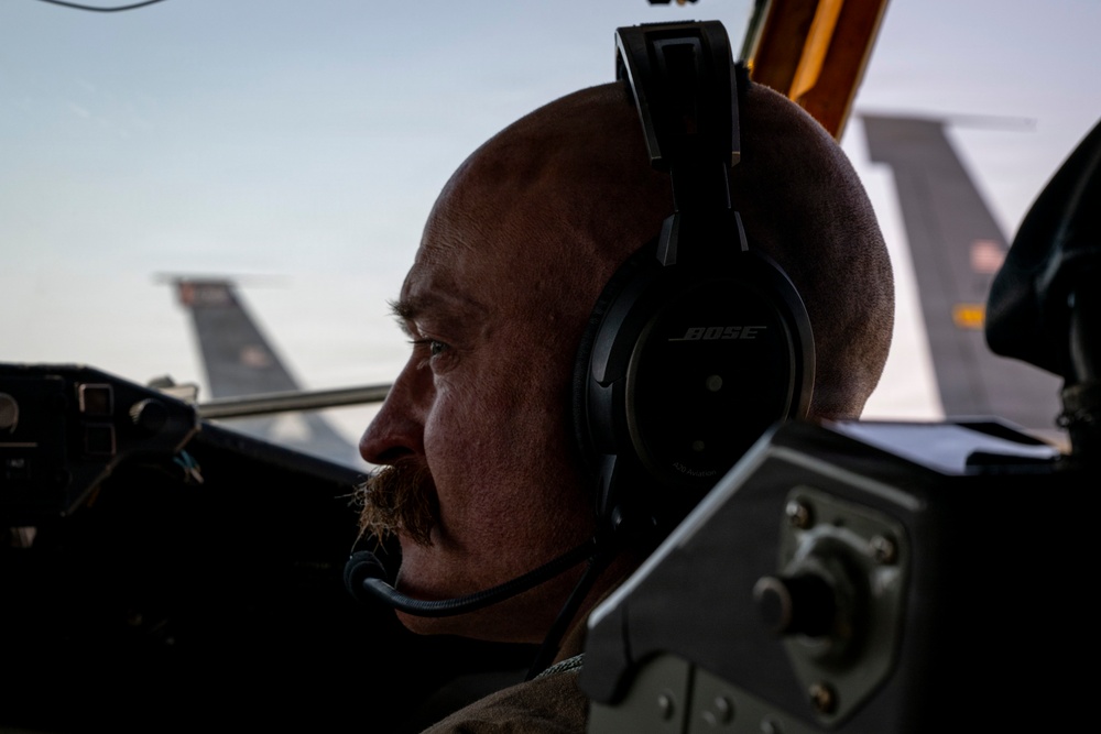 A bald man with a mustache wearing Bose aviation headphones with an attached microphone is seated in an aircraft cockpit. He is looking forward through the windscreen, with military aircraft tail sections visible in the background. The image is dramatically lit with strong contrast, suggesting early morning or late evening lighting conditions.