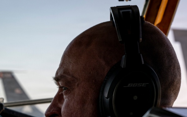 A bald man with a mustache wearing Bose aviation headphones with an attached microphone is seated in an aircraft cockpit. He is looking forward through the windscreen, with military aircraft tail sections visible in the background. The image is dramatically lit with strong contrast, suggesting early morning or late evening lighting conditions.