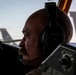 A bald man with a mustache wearing Bose aviation headphones with an attached microphone is seated in an aircraft cockpit. He is looking forward through the windscreen, with military aircraft tail sections visible in the background. The image is dramatically lit with strong contrast, suggesting early morning or late evening lighting conditions.