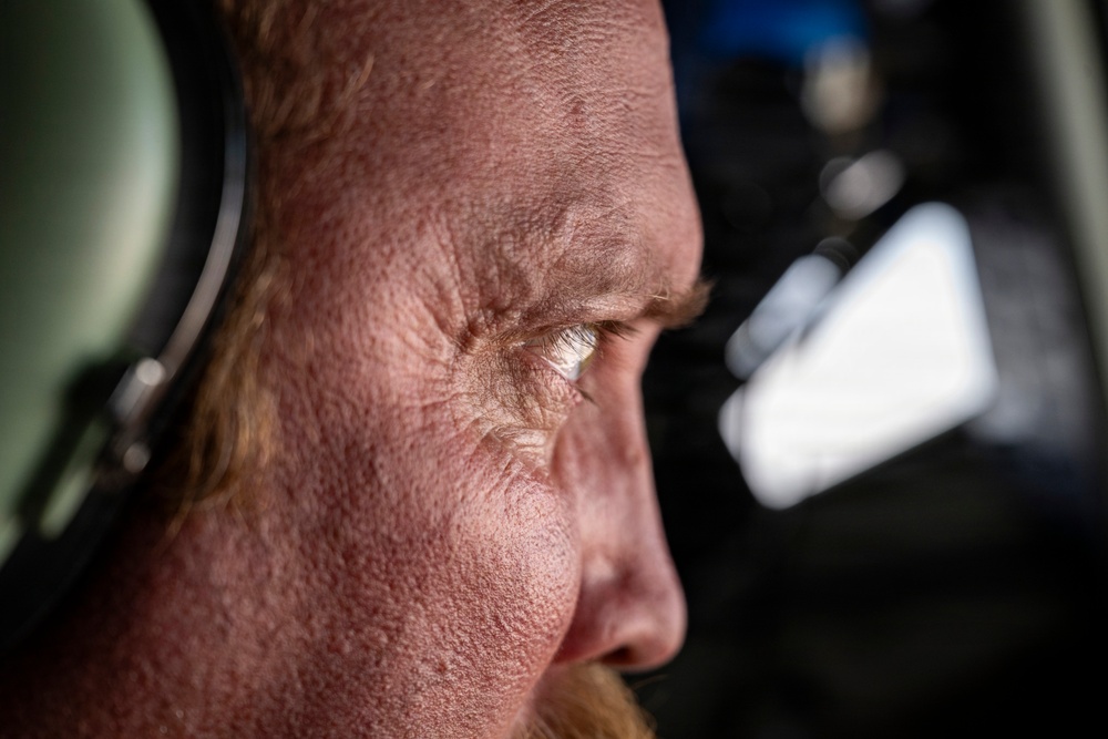 An extreme close-up profile shot of an older individual wearing aviation headphones, captured with shallow depth of field. The image reveals significant facial detail including skin texture, wrinkles, and reddish hair. The background suggests an aircraft cockpit or cabin environment.