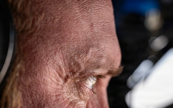 An extreme close-up profile shot of an older individual wearing aviation headphones, captured with shallow depth of field. The image reveals significant facial detail including skin texture, wrinkles, and reddish hair. The background suggests an aircraft cockpit or cabin environment.