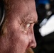 An extreme close-up profile shot of an older individual wearing aviation headphones, captured with shallow depth of field. The image reveals significant facial detail including skin texture, wrinkles, and reddish hair. The background suggests an aircraft cockpit or cabin environment.