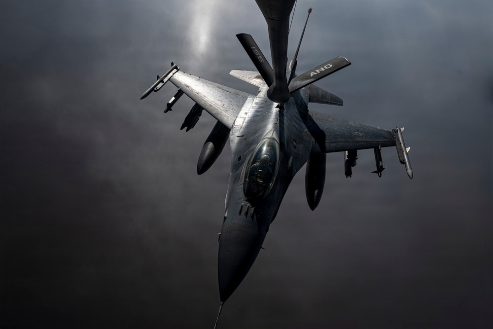 A U.S. Air National Guard (ANG) F-16 Fighting Falcon is captured in a dramatic aerial photograph during an in-flight refueling operation, visible from the refueling boom extending from above. The aircraft is armed with multiple air-to-air missiles and other ordnance on its wing pylons. The image is taken from the perspective of the tanker aircraft looking down at the fighter jet against a dark, overcast sky.