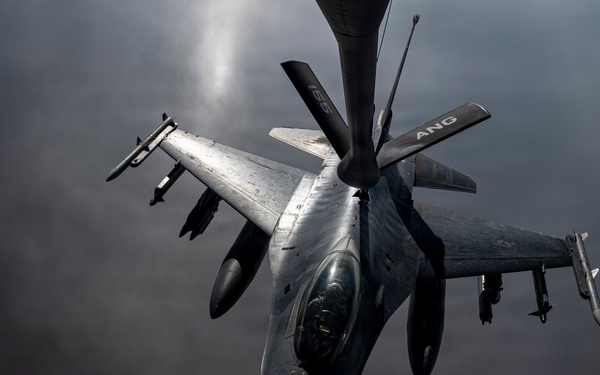 A U.S. Air National Guard (ANG) F-16 Fighting Falcon is captured in a dramatic aerial photograph during an in-flight refueling operation, visible from the refueling boom extending from above. The aircraft is armed with multiple air-to-air missiles and other ordnance on its wing pylons. The image is taken from the perspective of the tanker aircraft looking down at the fighter jet against a dark, overcast sky.