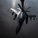 A U.S. Air National Guard (ANG) F-16 Fighting Falcon is captured in a dramatic aerial photograph during an in-flight refueling operation, visible from the refueling boom extending from above. The aircraft is armed with multiple air-to-air missiles and other ordnance on its wing pylons. The image is taken from the perspective of the tanker aircraft looking down at the fighter jet against a dark, overcast sky.