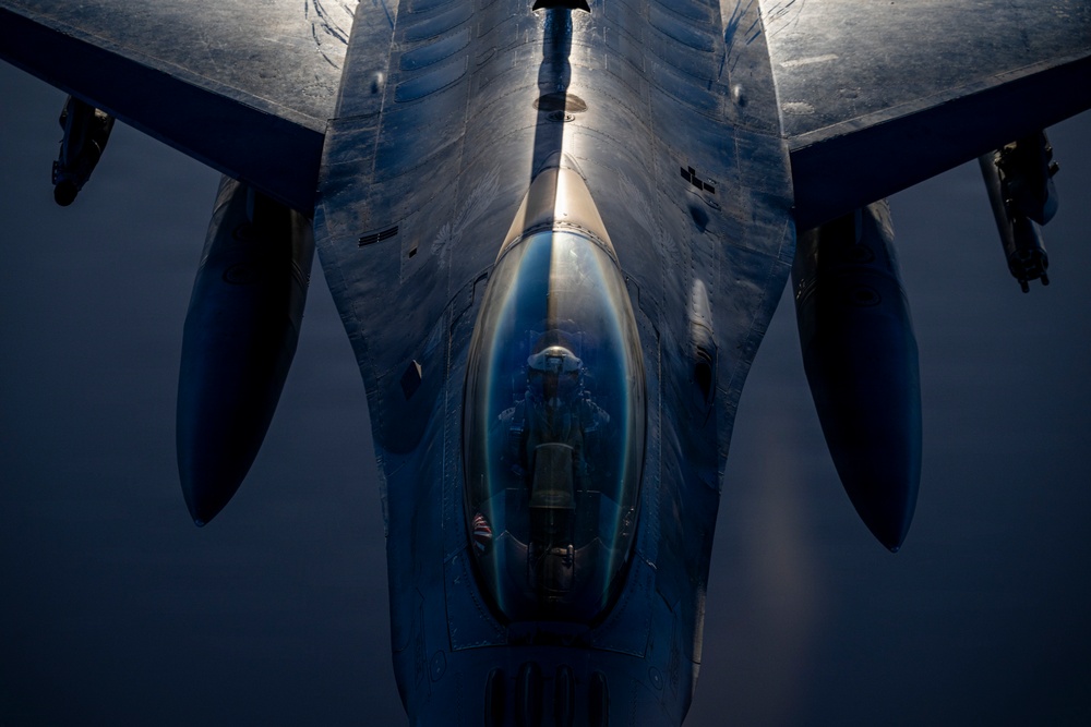 A close-up aerial photograph taken from above and behind a military fighter jet, likely an F-16 Fighting Falcon, captured during flight. The image is shot from a top-down perspective, possibly from a refueling tanker aircraft, revealing the cockpit canopy with a pilot visible inside. Dramatic lighting creates strong contrasts across the aircraft's metallic fuselage and delta wing configuration.