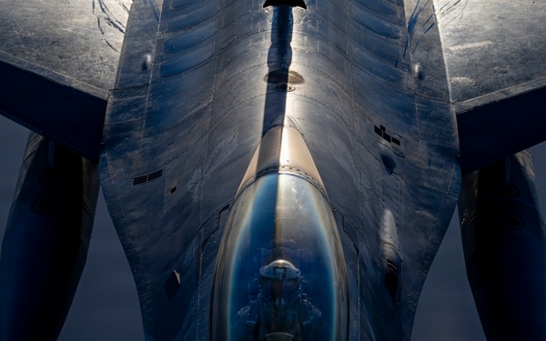 A close-up aerial photograph taken from above and behind a military fighter jet, likely an F-16 Fighting Falcon, captured during flight. The image is shot from a top-down perspective, possibly from a refueling tanker aircraft, revealing the cockpit canopy with a pilot visible inside. Dramatic lighting creates strong contrasts across the aircraft's metallic fuselage and delta wing configuration.