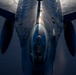 A close-up aerial photograph taken from above and behind a military fighter jet, likely an F-16 Fighting Falcon, captured during flight. The image is shot from a top-down perspective, possibly from a refueling tanker aircraft, revealing the cockpit canopy with a pilot visible inside. Dramatic lighting creates strong contrasts across the aircraft's metallic fuselage and delta wing configuration.