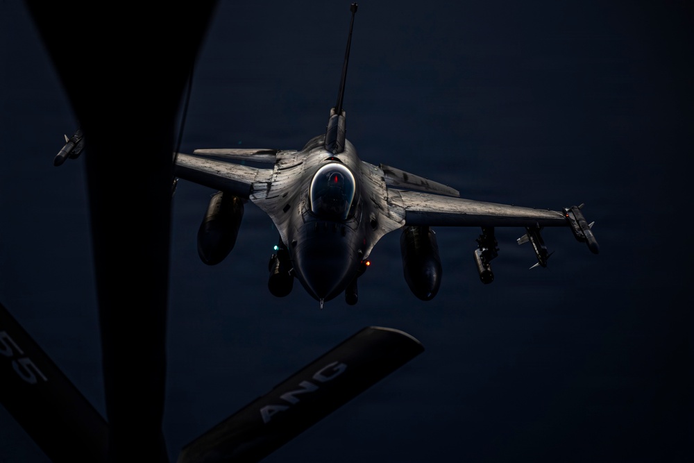 A fighter jet, identifiable as an F-16 Fighting Falcon, is captured from above during an aerial refueling operation at night or in low-light conditions. The image is taken from the perspective of a tanker aircraft, with the refueling boom visible in the foreground. The fighter is armed with multiple munitions on its wing hardpoints and is approaching the tanker for fuel transfer.