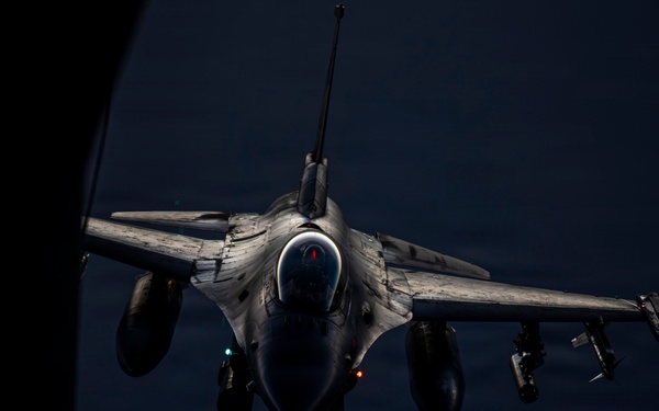 A fighter jet, identifiable as an F-16 Fighting Falcon, is captured from above during an aerial refueling operation at night or in low-light conditions. The image is taken from the perspective of a tanker aircraft, with the refueling boom visible in the foreground. The fighter is armed with multiple munitions on its wing hardpoints and is approaching the tanker for fuel transfer.