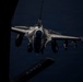 A fighter jet, identifiable as an F-16 Fighting Falcon, is captured from above during an aerial refueling operation at night or in low-light conditions. The image is taken from the perspective of a tanker aircraft, with the refueling boom visible in the foreground. The fighter is armed with multiple munitions on its wing hardpoints and is approaching the tanker for fuel transfer.