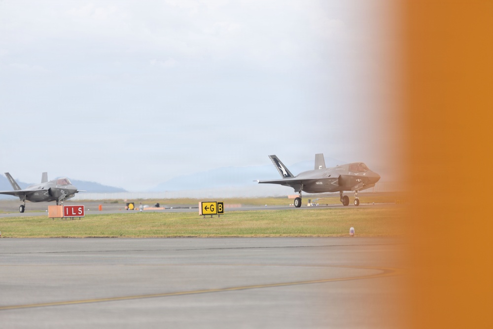 Two F-35 stealth fighter jets are visible on an airfield taxiway or apron, with one in the foreground and one in the background. Airfield signage including an 'ILS' marker (red) and a taxiway 'G B' directional sign (yellow) are clearly visible. The right portion of the image is obscured by an orange blur, likely caused by a nearby aircraft or object partially entering the camera frame.