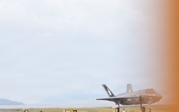 Two F-35 stealth fighter jets are visible on an airfield taxiway or apron, with one in the foreground and one in the background. Airfield signage including an 'ILS' marker (red) and a taxiway 'G B' directional sign (yellow) are clearly visible. The right portion of the image is obscured by an orange blur, likely caused by a nearby aircraft or object partially entering the camera frame.