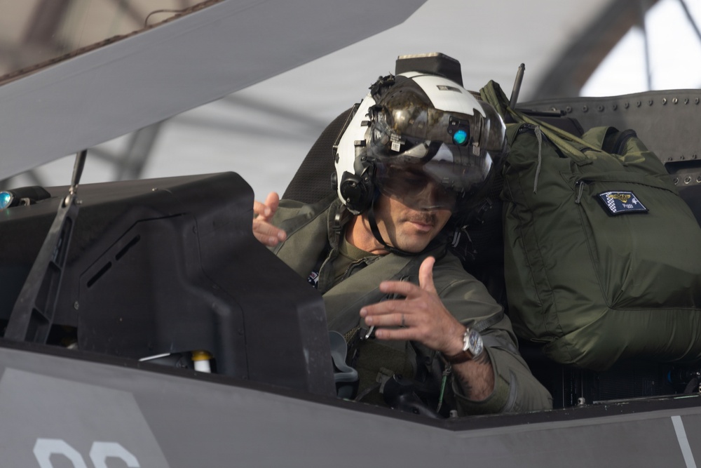 A military pilot is seated in the cockpit of what appears to be an F-35 stealth fighter jet. The pilot is wearing a sophisticated helmet-mounted display system with a visor and night vision attachment, along with an olive green flight suit. The pilot appears to be gesturing with both hands, possibly communicating with ground crew.