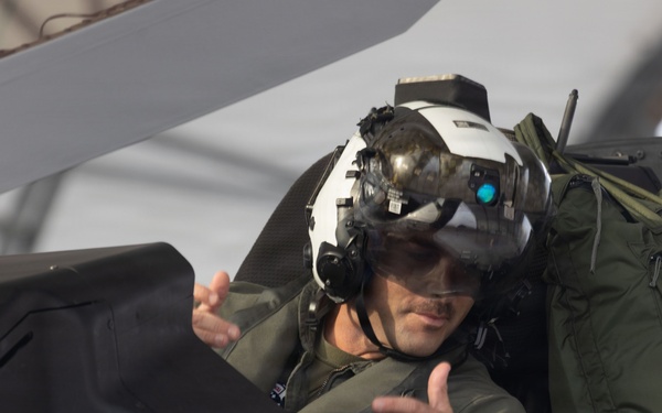 A military pilot is seated in the cockpit of what appears to be an F-35 stealth fighter jet. The pilot is wearing a sophisticated helmet-mounted display system with a visor and night vision attachment, along with an olive green flight suit. The pilot appears to be gesturing with both hands, possibly communicating with ground crew.