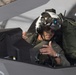 A military pilot is seated in the cockpit of what appears to be an F-35 stealth fighter jet. The pilot is wearing a sophisticated helmet-mounted display system with a visor and night vision attachment, along with an olive green flight suit. The pilot appears to be gesturing with both hands, possibly communicating with ground crew.