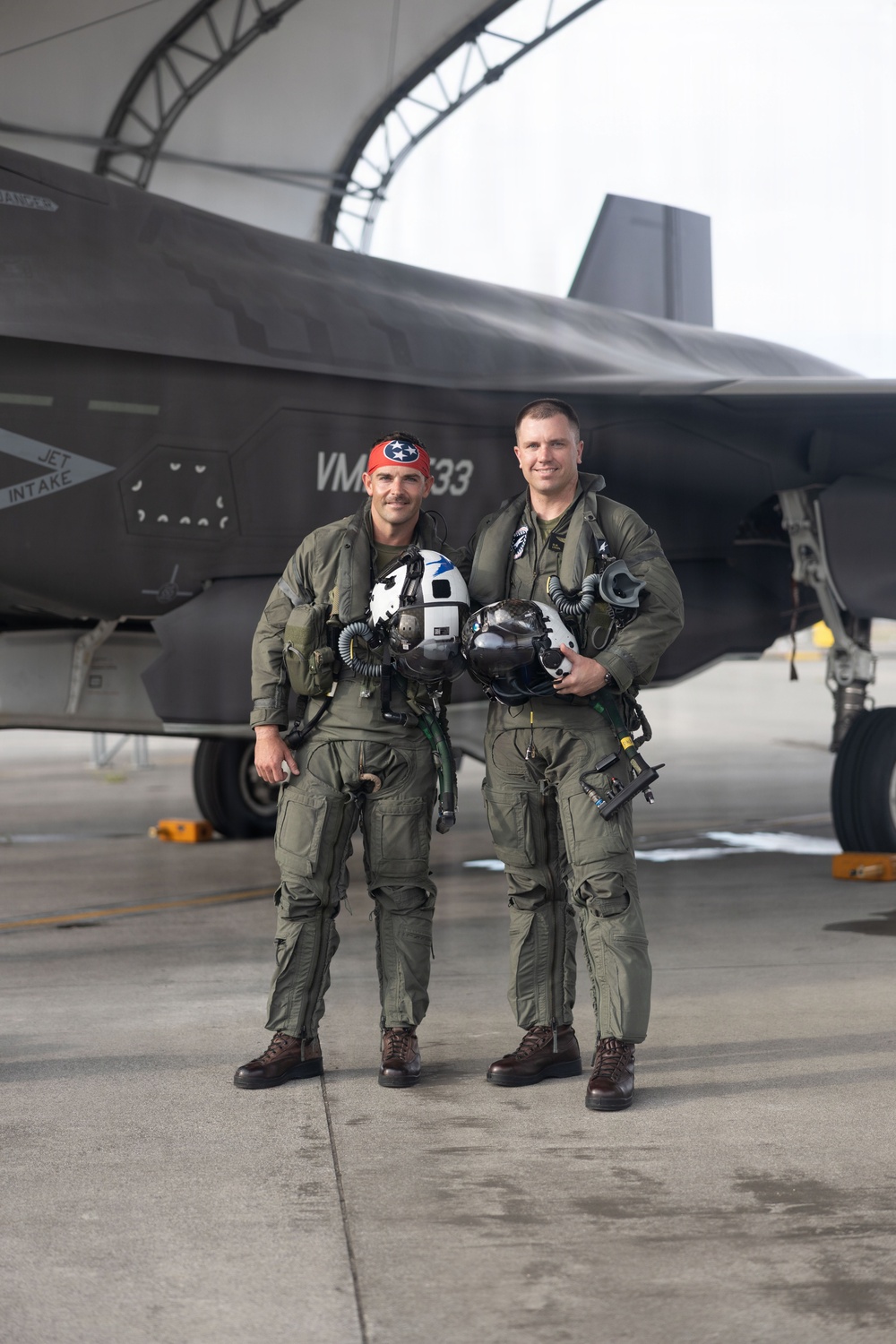 Two military pilots in olive green flight suits stand posed in front of a dark gray stealth fighter jet, likely an F-35B, under a curved aircraft hangar structure. Both are holding flight helmets and wearing full pilot gear including G-suits and survival equipment. The aircraft markings visible include 'VMA' and the number '33', along with a 'JET INTAKE' warning label.