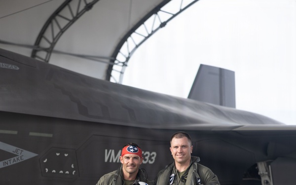 Two military pilots in olive green flight suits stand posed in front of a dark gray stealth fighter jet, likely an F-35B, under a curved aircraft hangar structure. Both are holding flight helmets and wearing full pilot gear including G-suits and survival equipment. The aircraft markings visible include 'VMA' and the number '33', along with a 'JET INTAKE' warning label.