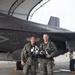 Two military pilots in olive green flight suits stand posed in front of a dark gray stealth fighter jet, likely an F-35B, under a curved aircraft hangar structure. Both are holding flight helmets and wearing full pilot gear including G-suits and survival equipment. The aircraft markings visible include 'VMA' and the number '33', along with a 'JET INTAKE' warning label.