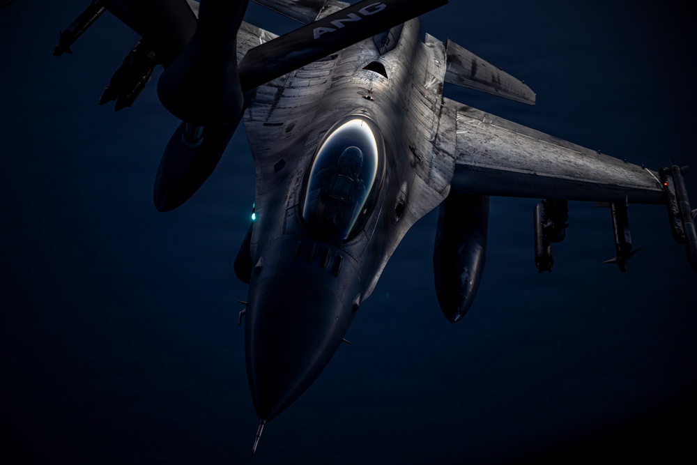A military fighter jet, identifiable as an F-16 Fighting Falcon, is photographed from above and slightly forward during what appears to be a nighttime aerial refueling operation. The image is taken from the perspective of a tanker aircraft, looking down at the receiving jet against a dark blue sky. The letters 'ANG' (Air National Guard) are partially visible on the tanker boom or fuselage above.
