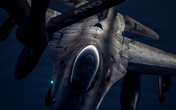 A military fighter jet, identifiable as an F-16 Fighting Falcon, is photographed from above and slightly forward during what appears to be a nighttime aerial refueling operation. The image is taken from the perspective of a tanker aircraft, looking down at the receiving jet against a dark blue sky. The letters 'ANG' (Air National Guard) are partially visible on the tanker boom or fuselage above.