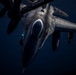 A military fighter jet, identifiable as an F-16 Fighting Falcon, is photographed from above and slightly forward during what appears to be a nighttime aerial refueling operation. The image is taken from the perspective of a tanker aircraft, looking down at the receiving jet against a dark blue sky. The letters 'ANG' (Air National Guard) are partially visible on the tanker boom or fuselage above.