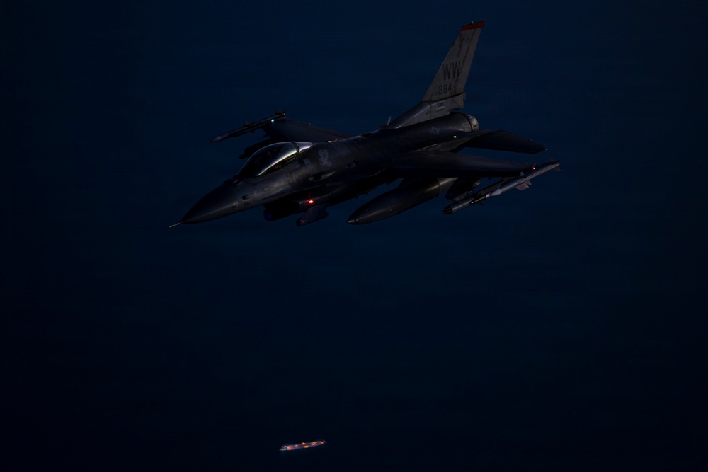 A U.S. Air Force F-16 Fighting Falcon is photographed in flight during nighttime conditions. The aircraft bears 'NW' tail code markings, indicating assignment to the 35th Fighter Wing based at Misawa Air Base, Japan. A munition or store appears to have just been released and is visible falling below the aircraft.