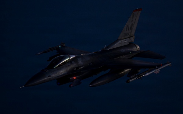 A U.S. Air Force F-16 Fighting Falcon is photographed in flight during nighttime conditions. The aircraft bears 'NW' tail code markings, indicating assignment to the 35th Fighter Wing based at Misawa Air Base, Japan. A munition or store appears to have just been released and is visible falling below the aircraft.