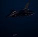 A U.S. Air Force F-16 Fighting Falcon is photographed in flight during nighttime conditions. The aircraft bears 'NW' tail code markings, indicating assignment to the 35th Fighter Wing based at Misawa Air Base, Japan. A munition or store appears to have just been released and is visible falling below the aircraft.