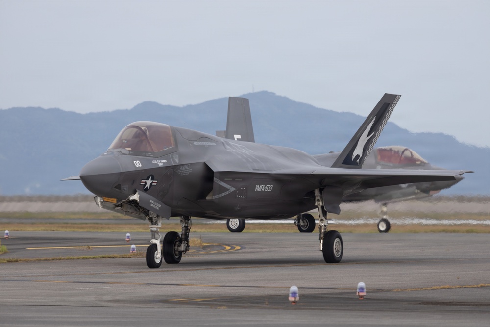 A U.S. Marine Corps F-35B Lightning II stealth fighter jet is taxiing on an airfield tarmac. The aircraft bears the markings of VMFA-533 squadron with tail code '00' and a distinctive shark/ghost tail art. A second aircraft is partially visible in the background to the right.