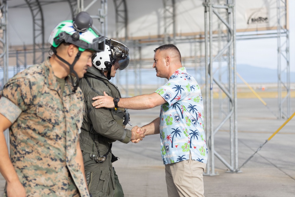 The image shows a greeting or welcome ceremony on a military airfield tarmac. A civilian in a tropical Hawaiian-style shirt is shaking hands with a military pilot wearing a flight helmet and olive green flight suit, while a U.S. Marine in MARPAT camouflage uniform stands nearby. The setting appears to be near a large aircraft hangar or maintenance structure.