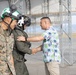 The image shows a greeting or welcome ceremony on a military airfield tarmac. A civilian in a tropical Hawaiian-style shirt is shaking hands with a military pilot wearing a flight helmet and olive green flight suit, while a U.S. Marine in MARPAT camouflage uniform stands nearby. The setting appears to be near a large aircraft hangar or maintenance structure.
