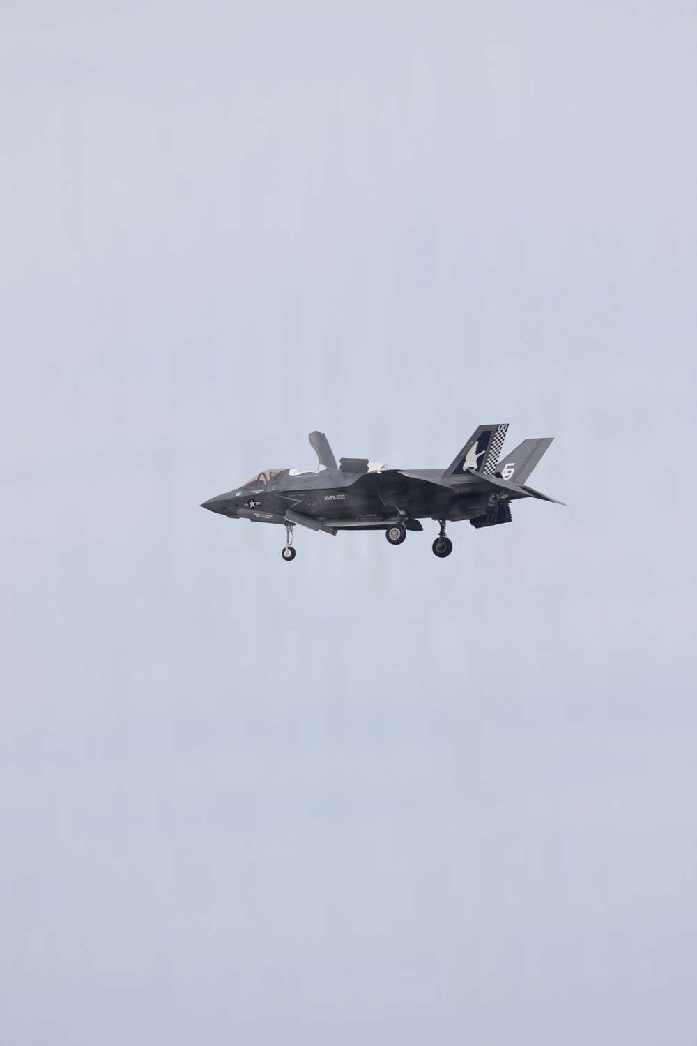 A single-seat military jet fighter is photographed in flight against an overcast gray sky. The aircraft has its landing gear deployed, suggesting it is in an approach or low-speed flight configuration. Markings and tail art indicate it is a U.S. Marine Corps aircraft.