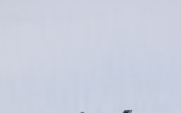 A single-seat military jet fighter is photographed in flight against an overcast gray sky. The aircraft has its landing gear deployed, suggesting it is in an approach or low-speed flight configuration. Markings and tail art indicate it is a U.S. Marine Corps aircraft.